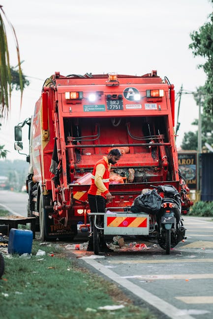 A worker wearing a red and yellow uniform is seen operating a red waste collection truck on a roadside, with the truck’s rear compartment open and partially filled with waste material. The worker is leaning slightly forward, handling or inspecting the trash, which is loaded into the rear of the vehicle. In front of the truck, there is a black rubbish bag placed on a small stand or platform attached to a motorbike parked nearby. The environment includes a paved roadside with a curb, some green grass, and scattered debris on the ground. In the background, there are trees, utility poles with wires, and buildings, indicating an urban or suburban setting. The scene appears to be during daytime with natural light illuminating the area. The image aligns with the context of independent waste collection and rubbish removal services, akin to those offered by Waste Removal Greenwich, highlighting on-site clearance and waste management activities on a residential or commercial street.