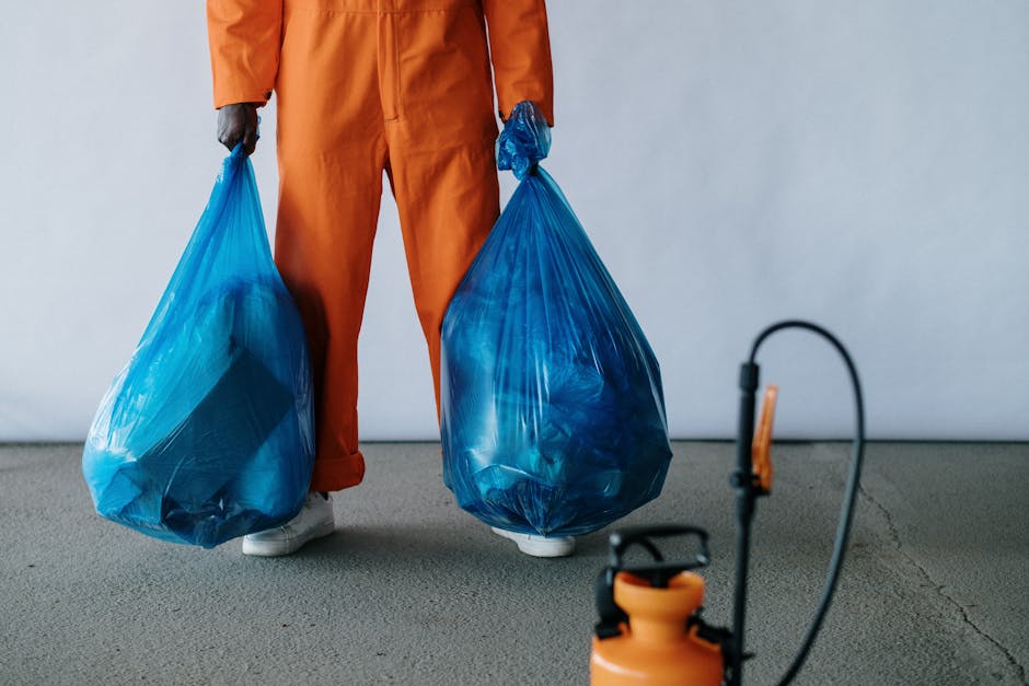 A person dressed in orange work overalls and white shoes is holding two large blue plastic bags filled with rubbish, one in each hand. The bags appear to be semi-transparent, revealing mixed waste inside. The individual is standing indoors on a concrete surface with a plain, light-colored background. In the foreground, part of an orange waste sprayer or pump is visible, along with a black tube connected to it, suggesting a setting related to waste management or rubbish collection. The scene emphasizes the process of collection and handling of waste materials, aligning with private rubbish removal or alternative on-site clearance services offered by Waste Removal Greenwich. The lighting is neutral, and the environment appears clean and organized, with no other visible objects or clutter, highlighting the professional approach to waste disposal.