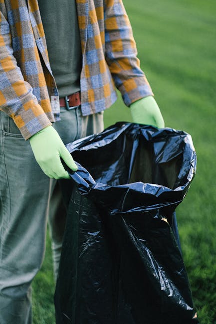 A worker wearing a red and yellow uniform is seen operating a red waste collection truck on a roadside, with the truck’s rear compartment open and partially filled with waste material. The worker is leaning slightly forward, handling or inspecting the trash, which is loaded into the rear of the vehicle. In front of the truck, there is a black rubbish bag placed on a small stand or platform attached to a motorbike parked nearby. The environment includes a paved roadside with a curb, some green grass, and scattered debris on the ground. In the background, there are trees, utility poles with wires, and buildings, indicating an urban or suburban setting. The scene appears to be during daytime with natural light illuminating the area. The image aligns with the context of independent waste collection and rubbish removal services, akin to those offered by Waste Removal Greenwich, highlighting on-site clearance and waste management activities on a residential or commercial street.