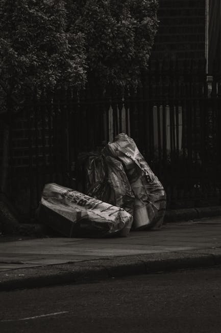 On a paved street curb in an outdoor urban environment, three large black garbage bags made of thick plastic are placed side by side, leaning against a dark wooden fence. The bags appear to be filled with waste material, with one slightly crumpled and the others more tightly compressed. Behind the fence, dense foliage from trees or shrubs provides a leafy background, casting partial shadows over the scene. The pavement in the foreground is smooth with a visible white line marking the edge of the road, and the overall lighting suggests an overcast or shaded setting. The scene reflects an area where private waste disposal or rubbish collection services, such as those offered by Waste Removal Greenwich, might be used to handle bulky refuse outside standard council collection services, illustrating typical on-site clearance or independent rubbish removal practices.