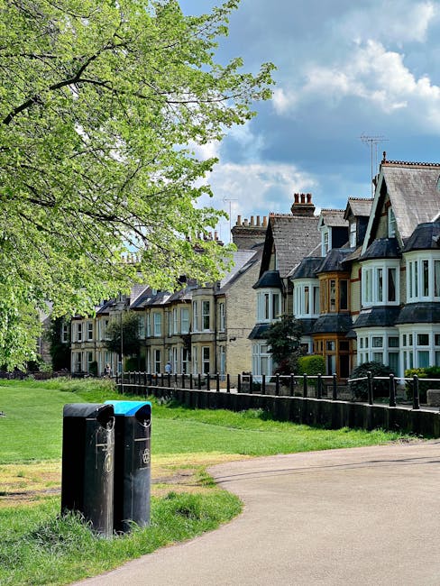 In a residential neighborhood, two black waste bins are positioned on a grassy verge adjacent to a paved footpath. Behind the bins, a row of traditional Victorian-style terraced houses with brick facades, bay windows, and varied rooflines can be seen, with some featuring dormer windows and chimneys. To the left, a large deciduous tree with fresh green leaves extends its branches over the scene, providing partial shade and a natural backdrop. The sky above is mostly clear with some scattered clouds, suggesting a bright day. The area appears tidy and well-maintained, with a black wooden fence separating the front gardens of the houses from the green public space. This scene captures an everyday aspect of urban waste management, where independent collection points such as these are used for rubbish collection, offering an alternative to council bin services, and is relevant to private waste handling in residential areas.