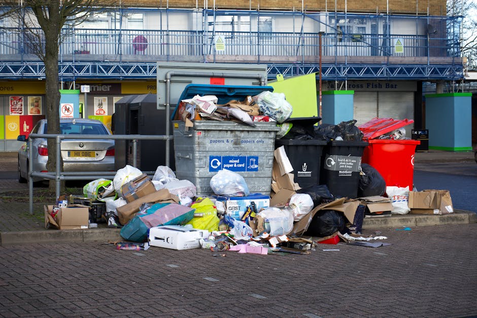 A large collection of overflowing waste and rubbish bags, including black, white, and transparent plastic bags, cardboard boxes, and paper packaging, all piled in front of a grey communal bin with a blue lid, situated on a raised concrete platform at the edge of a paved parking area. The rubbish consists of discarded packaging, flattened boxes, and various paper and plastic items. Behind the waste are parked vehicles, including a silver car with the registration plate partially visible, and in the background, a building with a blue metal scaffolding structure, storefronts, and signs. The scene indicates a scenario of uncollected rubbish, possibly related to private waste management services. The environment features a paved area with a metal railing on the left side, a tree trunk also on the left, and the overall setting appears to be an urban or commercial area in need of rubbish removal services, highlighting the importance of timely and proper waste disposal.