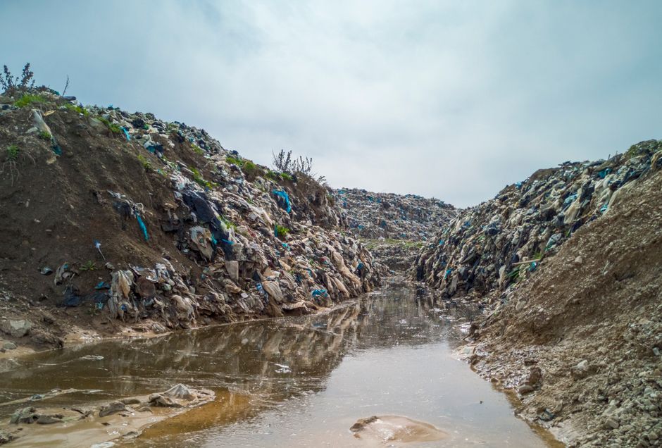 A narrow, exposed trench filled with stagnant, muddy water, with steep, uneven sides composed of soil and loose debris. The sides are heavily littered with various types of waste, including plastic bags, bottles, and fragments of fabric and paper, with some items deeply embedded into the soil. The water appears brown and murky, reflecting the overcast sky above, which is partially visible in the background. Sparse, small patches of grass and weeds grow on the top edges of the trench, which extends into the distance, suggesting an area affected by illegal or neglected waste dumping. The environment indicates neglect and contamination, supporting the context of waste accumulation and the need for proper rubbish removal or disposal services. This scene illustrates the consequences of improper waste management and the importance of regular clearance, such as that offered by Waste Removal Greenwich, to prevent such environmental issues.