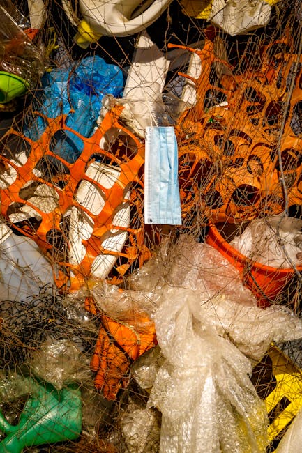 A large collection of overflowing waste and rubbish bags, including black, white, and transparent plastic bags, cardboard boxes, and paper packaging, all piled in front of a grey communal bin with a blue lid, situated on a raised concrete platform at the edge of a paved parking area. The rubbish consists of discarded packaging, flattened boxes, and various paper and plastic items. Behind the waste are parked vehicles, including a silver car with the registration plate partially visible, and in the background, a building with a blue metal scaffolding structure, storefronts, and signs. The scene indicates a scenario of uncollected rubbish, possibly related to private waste management services. The environment features a paved area with a metal railing on the left side, a tree trunk also on the left, and the overall setting appears to be an urban or commercial area in need of rubbish removal services, highlighting the importance of timely and proper waste disposal.