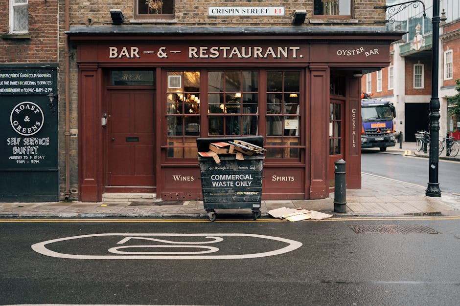 In front of a red-brick building housing a bar and restaurant, there is a large black commercial waste bin positioned on the pavement. The bin is made of durable plastic with a hinged lid and is marked with white lettering indicating it is designated for commercial waste only. It is partially overflowing with flattened cardboard boxes and other paper packaging, with some cardboard pieces resting on the ground nearby. The on-street scene includes a white circular road marking with a line through the center, and a black bollard is situated to the right of the bin, preventing vehicle access to the sidewalk. To the left, a black signboard advertises a self-service buffet, while a black lamppost is located further to the right. In the background, a parked truck and bicycles can be seen at the corner, suggesting this area caters to local businesses and pedestrians. The scene emphasizes on-site waste management and the importance of proper rubbish handling in urban commercial settings, with Waste Removal Greenwich providing tailored solutions for such environments.