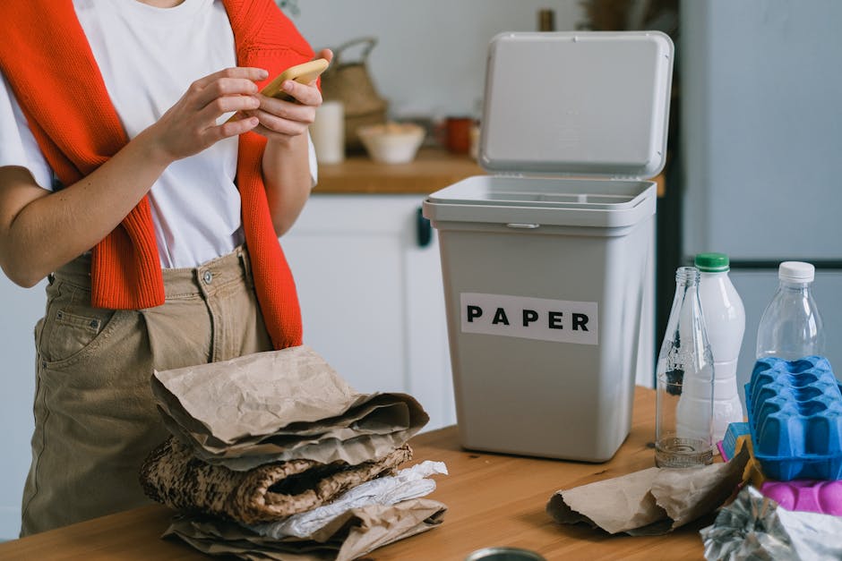 A person standing in a kitchen holding a mobile phone, with a pile of crumpled paper and brown paper bags on the wooden countertop in front. To the right, there is a white domestic waste bin labeled 'PAPER' in bold black letters, with the lid open. Near the bin, there are clear plastic bottles, some with caps, and a blue plastic egg tray. The background shows a blurred kitchen environment with a white cabinet, some bowls, and decorative items. The scene is well-lit with natural or ambient lighting, emphasizing the importance of correct waste sorting for recycling, and illustrating the use of a dedicated paper bin as part of private waste disposal practices in household recycling setups, aligning with waste removal services.