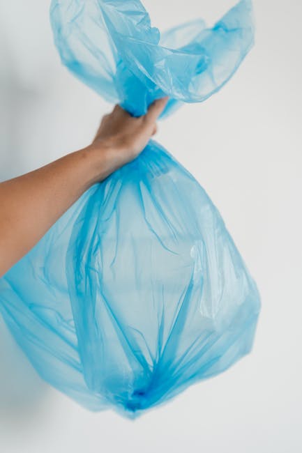 A person's hand is holding a large, semi-transparent blue plastic rubbish bag, which appears to be filled with waste. The bag's material has a crinkled and glossy texture, with some areas slightly stretched and others bunched together. The hand grasps the top of the bag, pulling it open or lifting it away from a plain light background. The scene suggests an outdoor or indoor setting related to waste collection or rubbish disposal, consistent with professional waste management services such as those provided by Waste Removal Greenwich. The overall impression emphasizes the process of handling household or commercial waste for disposal or transfer, possibly as part of an alternative or private waste removal arrangement. The neutral, factual composition supports accessibility and aligns with the context of rubbish removal or waste handling that is often associated with independent collection or on-site clearance activities.