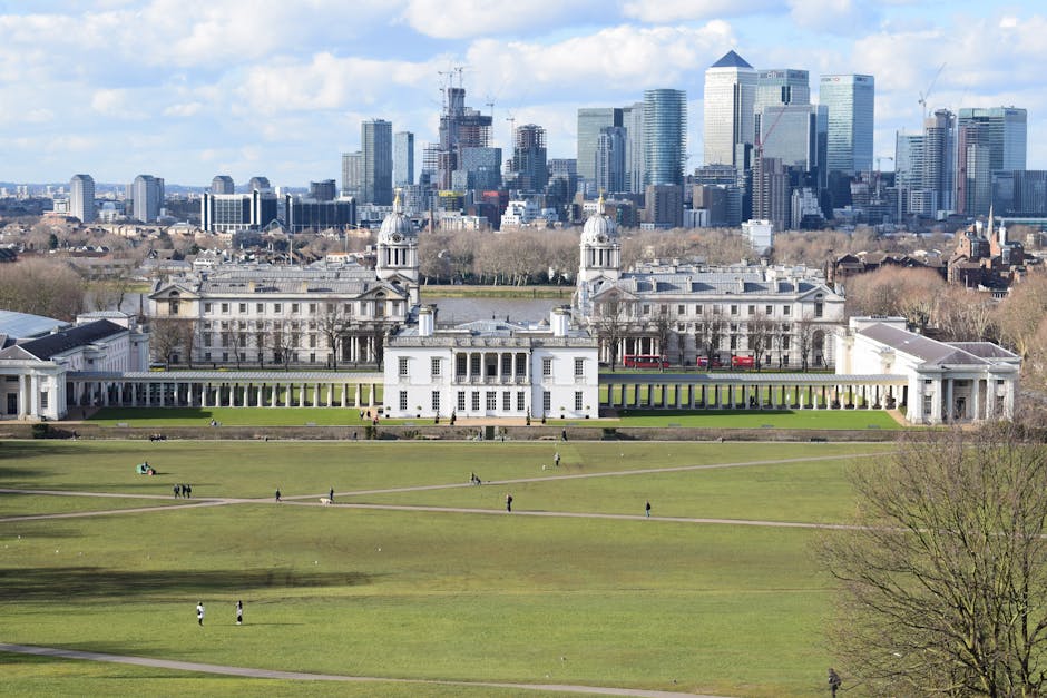 A panoramic view of a large, historic white palace with classical architectural features, including symmetrical columns, arched windows, and a central entrance, situated in a landscaped park with expansive green lawns and a few visitors walking along paved pathways. The palace is flanked by long wings extending outward, with small cupola-topped towers visible on either side. In the background, a modern city skyline with numerous high-rise office buildings, some under construction with cranes, rises against a partly cloudy sky, creating a stark contrast between historic and contemporary architecture. The foreground shows a well-maintained grassy park area with scattered pedestrians, some sitting on benches or walking, and a few trees with budding leaves suggesting early spring. The scene is lit with natural daylight, highlighting the details of the historic structure and the surrounding environment, subtly illustrating the theme of private or alternative disposal environments versus public waste management methods, relevant to rubbish removal services such as Waste Removal Greenwich.