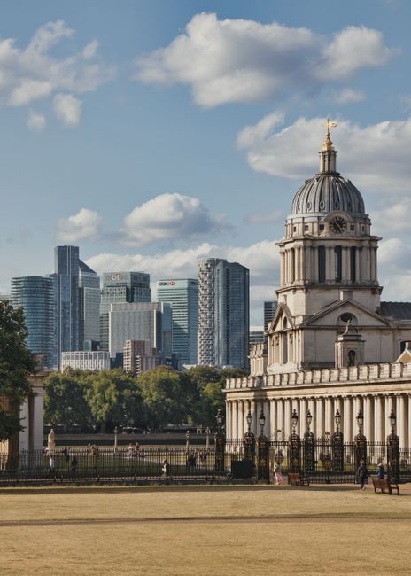 A historic stone building with a prominent domed clock tower featuring ornate architectural details and a golden weather vane at the top, situated in a landscaped park with a low iron fence surrounding it. In the background, a modern city skyline with tall glass office buildings and skyscrapers under a partly cloudy sky is visible, highlighting the contrast between historic and contemporary architecture. The foreground shows a well-maintained lawn with a few benches and several small groups of people walking or sitting, indicating a public space used for leisure and possibly for organising private waste collection or on-site clearance by waste removal services. The overall scene captures an urban environment where traditional and modern elements coexist, relevant to alternative waste handling options such as rubbish removal services by Waste Removal Greenwich.