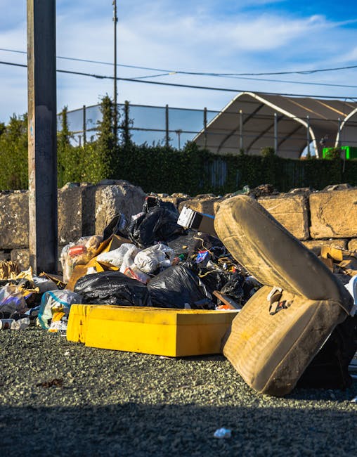 A close-up view of a mixed pile of waste spilling onto a gravel surface beside an old, partially dislodged tyre and a yellow plastic box, with several tightly packed black and white rubbish bags, some torn open revealing scattered scraps of paper, plastic, and other debris. The waste is situated outdoors near a stone wall composed of rough, uneven blocks, with a metal utility pole and overhead wires visible to the left. In the background, there is a hedge, a large tent-like structure with a curved roof, and a clear blue sky with a few wispy clouds. The scene suggests an unauthorised or unmanaged waste accumulation site, typical of an area where independent rubbish disposal or collection services, such as those offered by Waste Removal Greenwich, are used for non-standard waste clearance purposes.