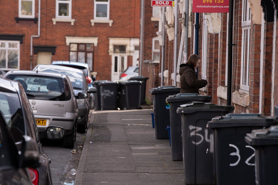 A narrow residential street in Greenwich lined with red brick terraced houses and parked vehicles on the left side, including a silver compact car and a darker hatchback. The pavement on the right side features a series of large, black wheelie bins with white spray-painted numbers on their sides, arranged in a row along the front of the houses. A woman wearing a brown coat is seen standing next to one of the bins, opening or closing the lid. The background includes additional parked cars and the continuation of the row of buildings, with a slightly overcast sky and natural daylight illuminating the scene. The setting emphasizes the common arrangement of private rubbish disposal containers placed outside homes, reflecting aspects of alternative waste management and independent collection methods facilitated by waste removal services such as Waste Removal Greenwich.