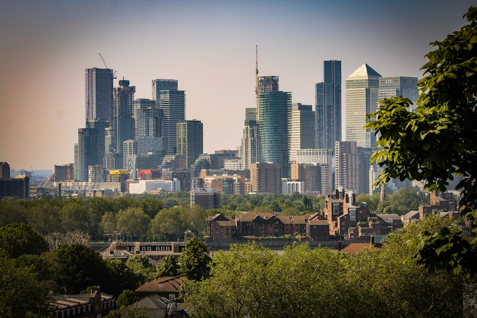 A collection of large skyscrapers and modern office buildings in the city skyline, with reflective glass facades in shades of blue and grey, towering over lower residential brick houses and green trees in the foreground. The buildings vary in height and architectural design, some topped with cranes indicating ongoing construction. The scene is illuminated by natural daylight, with clear skies above. The foreground features leafy green foliage and rooftops, suggesting an urban environment blending residential and commercial areas. This urban landscape emphasizes the scale and diversity of city structures, relevant to waste management services like those offered by Waste Removal Greenwich, which may involve on-site clearance or private disposal in complex city settings, especially involving high-rise and commercial waste collection.
