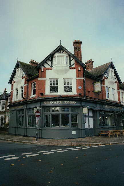 A traditional corner pub building with a distinctive Tudor-style architectural design featuring exposed black timber framing and red-brick walls, topped with a pitched slate roof and multiple chimneys. The ground floor is painted in a dark grey or navy color, contrasting with the white upper sections, and has large display windows behind which interior lighting is visible. The pub's signage reads 'GREENWICH TAVERN' in gold letters. The building is situated at the intersection of two quiet streets, with a sidewalk, a parked bollard, and a wooden bench on the pavement. The environment appears overcast with natural daylight providing even illumination, and minimal surrounding elements suggest a residential or commercial area in Greenwich. The scene relates to waste management services such as rubbish collection, which might be provided for businesses or local residents, emphasizing the importance of proper waste handling associated with pub and commercial properties. The photo is captured from ground level, giving a clear view of the building’s exterior details and surroundings.